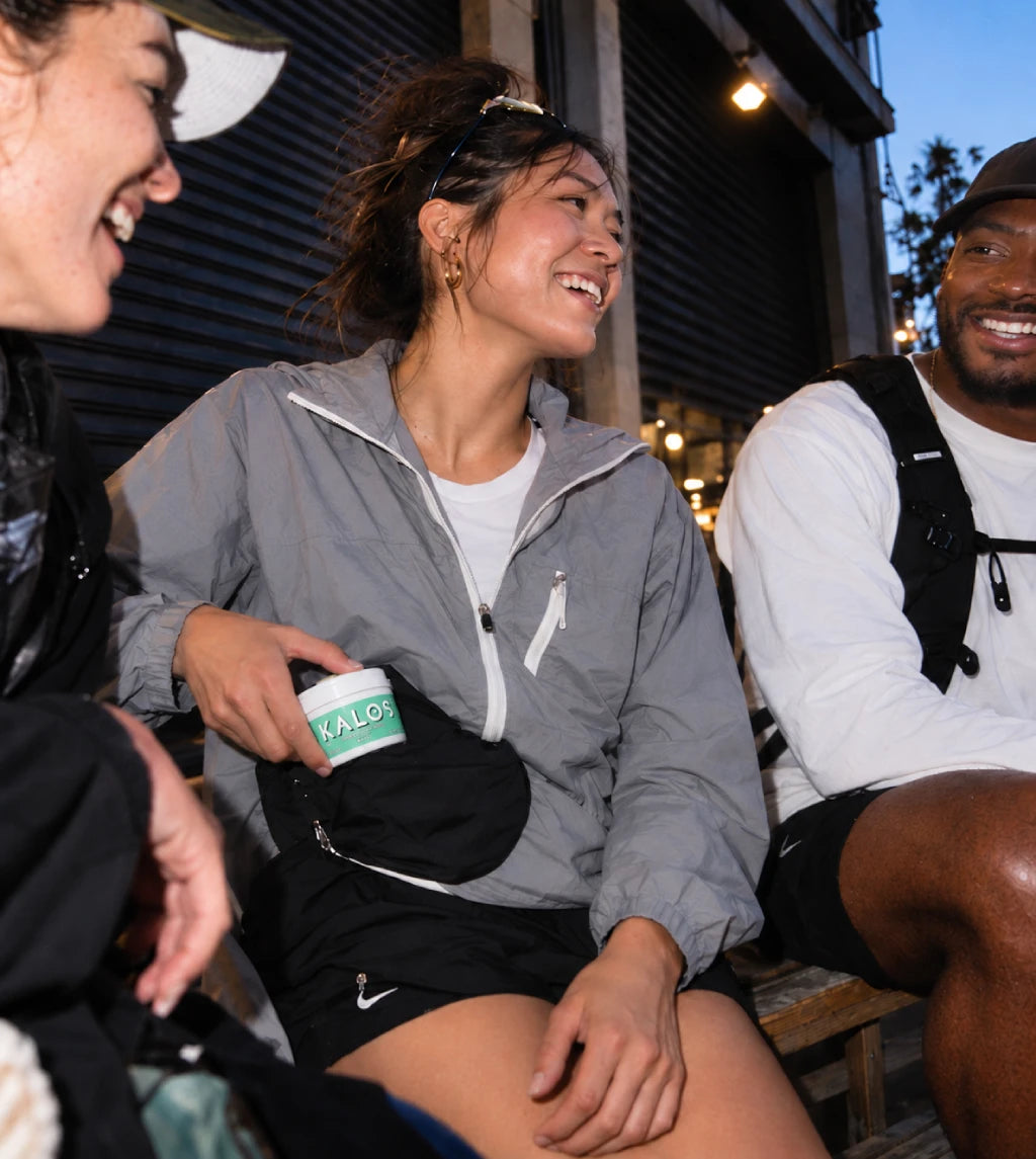A woman in a gray zip-up jacket and black shorts sits outdoors smiling, holding a small white jar with a green label that reads KALO. Two other people are partially visible at the sides in a casual evening setting.
